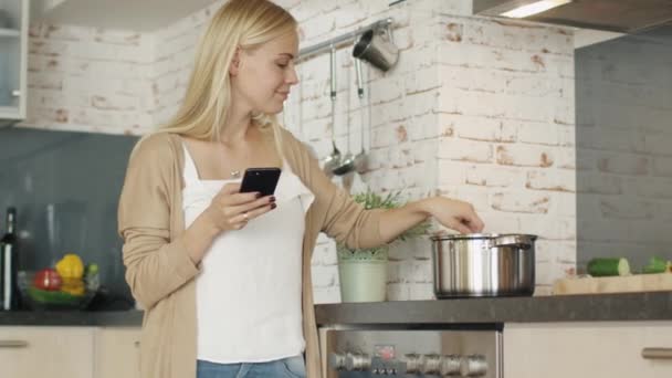 Pleasant Woman fica na cozinha antes do fogão enquanto usa seu smartphone. Ela agita algo na panela . — Vídeo de Stock