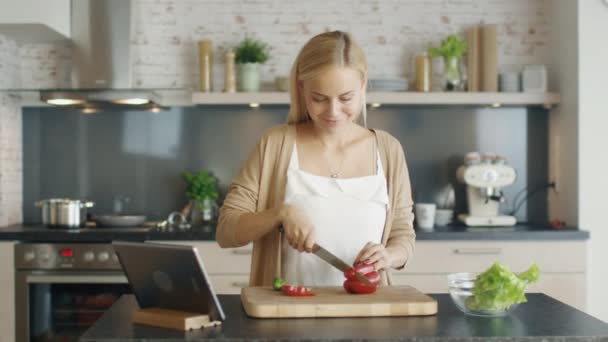 Heerlijke glimlach Blonde snijdt groenten op de keukentafel, vóór haar zit Tablet PC. — Stockvideo