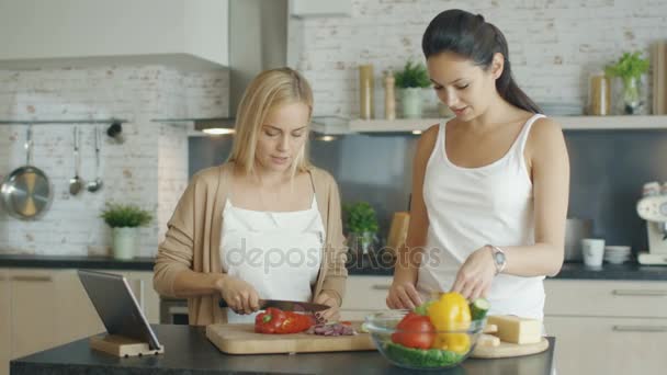 Zwei charmante Mädchen bereiten Salat zu, ein Tablet-Computer steht vor ihnen auf dem Tisch. Auch auf dem Herd wird gekocht. — Stockvideo