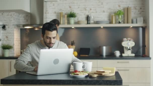 Na cozinha jovem Senta-se à mesa Laptop diante dele, Ele chama sua linda namorada e mostra-lhe algo na tela. Ambos riem. . — Vídeo de Stock