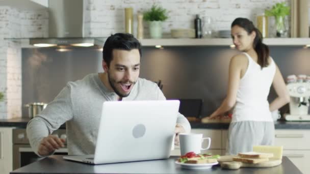 Assis dans la cuisine avant ordinateur portable jeune homme a de bonnes nouvelles et appelle sa petite amie qui cuisine. Les deux rient et sont heureux . — Video