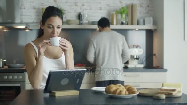 Vacker ung kvinna sitter vid bordet i köket medan du använder Tablet PC: N, klunkar kaffe och äter croissanter. Ung Man i bakgrunden kockar. — Stockvideo