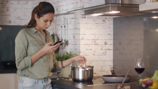 Молодая женщина перемешивает еду в кастрюле, держа свой смартфон и улыбаясь . — стоковое видео