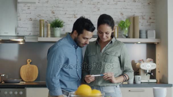 En la cocina hermosa chica muestra el resultado de la prueba de embarazo a su novio. Ambos abrazan felizmente. Moción lenta . — Vídeo de stock