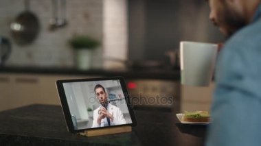 Genç adam danışır Online doktor tarafından mutfak masasında otururken kullanarak Tablet.