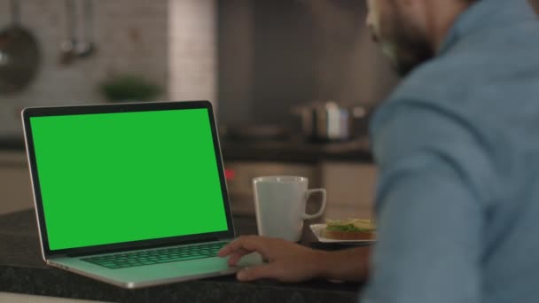 Junger Mann benutzt Laptop mit grünem Bildschirm während er am Küchentisch sitzt. — Stockvideo