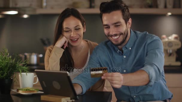 Casal feliz usa tablet enquanto sentado na mesa da cozinha. Homem detém cartão de crédito em sua mão . — Vídeo de Stock