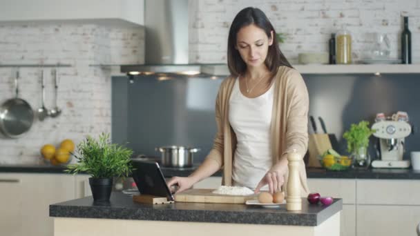 젊은 여자는 부엌에서 요리. 그녀는 그녀의 태블릿 컴퓨터와 나누기 측정된 밀가루의 힙으로 달걀에 레시피 참조. — 비디오