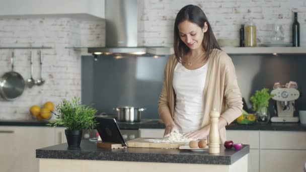 Genç kız un yemek pişirmek için hazırlar. Tarifi başvuru Tablet standları için mutfak masasının üzerinde. Yakışıklı işadamı sevgilisi ona gelir ve Hugs. Her ikisi de gülümseme. — Stok video