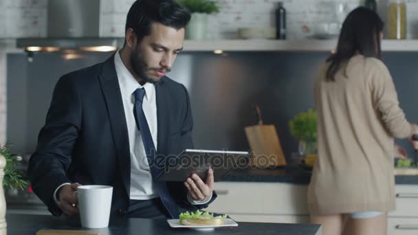 Hombre de negocios guapo utiliza Tablet Computer mientras desayuna en su cocina mientras su novia cocina en el fondo . — Vídeos de Stock