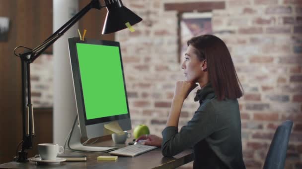 美しい女性は、思慮深く彼女のスタイリッシュなオフィスに坐っている間彼女のコンピューターで動作します。デスクトップ コンピューターは、緑色の画面を示しています。. — ストック動画