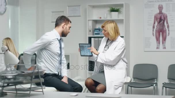 Mid Adult Female Doctor Consults Young Man About His Back Pain. Doctor le muestra Tablet Computer con su columna vertebral de rayos X . — Vídeos de Stock