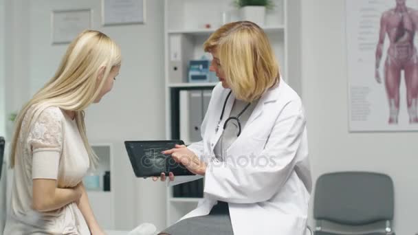 Médico femenino adulto medio consulta a mujer joven mostrando a su computadora de la tableta con la imagen del ultrasonido. La mujer sostiene su estómago. Ambos sonrían . — Vídeo de stock