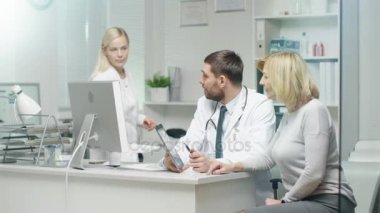 Profesyonel doktor orta yetişkin kadın danışır. O onun için ekrandaki bir dikkat çekiyor. Hemşire belgeleri doktor masanın üzerine koyar..