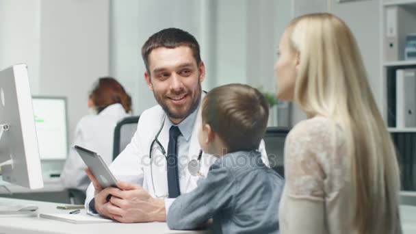 Médico Masculino Consulta Jovem Menino e Sua Mãe. Ele mostra algo em um computador tablet para eles . — Vídeo de Stock