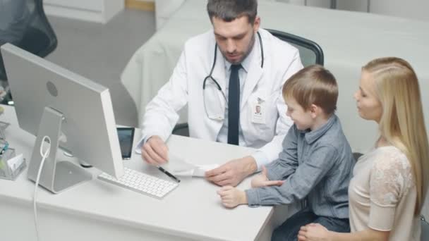 Durante la consulta médica, el médico masculino escribe una receta médica a un niño pequeño y se la da a su madre . — Vídeos de Stock