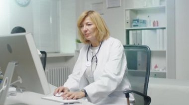 Doktorun ofisinde. Yetişkin kadın doktor masaüstü bilgisayar başında çalışıyor. Yardımcısı ayağa kalkar ve halk gösterilerden.