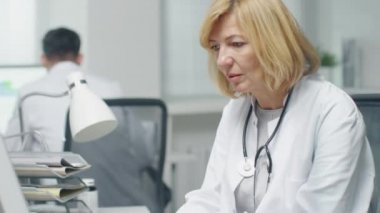 Doktorun ofisinde. Yetişkin kadın doktor masaüstü bilgisayar başında çalışıyor. Arka planda Yardımcısı masasında çalışıyor..