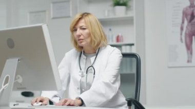 Doktorun ofisinde. Yetişkin kadın doktor masaüstü bilgisayar başında çalışıyor. Arka planda Yardımcısı masasında çalışıyor..