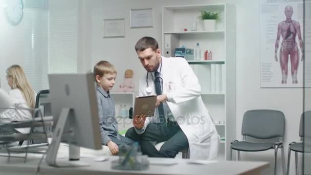 Médico Masculino Consulta Jovem Menino, Mostrando-lhe Tablet. Eles sorriem e brincam calorosamente . — Vídeo de Stock