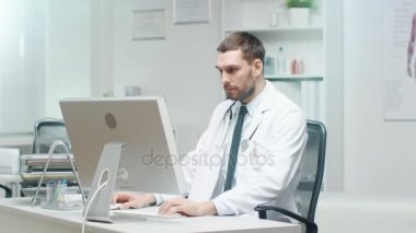 Erkek doktor onun masasında çalışıyor. Kişisel bilgisayar kullanır ve danışır belgeleri.