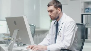 Erkek doktor onun masasında çalışıyor. Kişisel bilgisayar kullanır ve danışır belgeleri.
