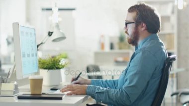 Genç sakallı adam alarak masaüstü bilgisayarda çalışırken notları. Kahve Sips. Yaratıcı ressam ofisinde oturuyor.