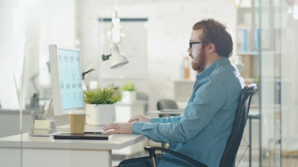 Молодой бородатый человек, работающий за персональным компьютером, сидя в офисе своего творческого агентства . — стоковое видео