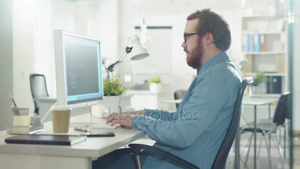 Brodaty kreatywny człowiek działa na kod, siedząc przy biurku. Jego siedziba znajduje się światła i nowoczesne. — Wideo stockowe
