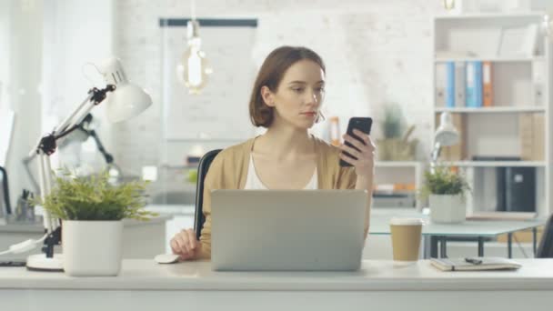 Retrato de una mujer creativa sentada en su escritorio. Ella sostiene el teléfono inteligente y consulta su cuaderno. Ella se sienta en una oficina ligera y moderna . — Vídeos de Stock