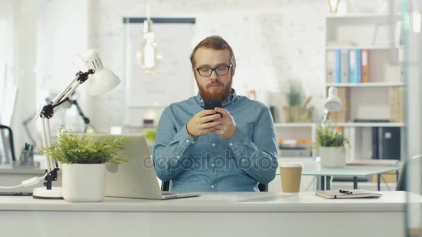 Retrato Tiro de um jovem barbudo sentado em sua mesa usando Smarphone. Notebook aberto mentiras sobre a mesa, escritório é leve e moderno . — Vídeo de Stock