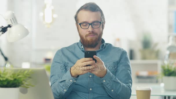 Portret van een jonge, bebaarde Man met bril zit aan zijn Bureau in een heldere kantoor via Smartphone. — Stockvideo
