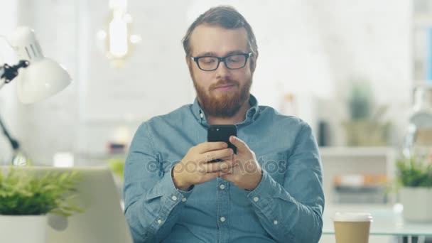 Portret młodego Brodaty mężczyzna w okularach siedzący przy biurku w biurze jasne za pomocą smartfona. — Wideo stockowe