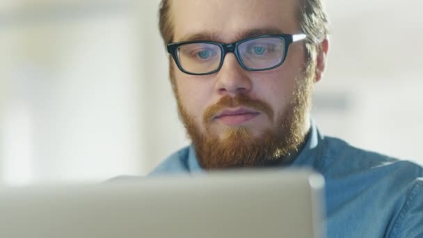 Portret van een bebaarde jonge Man met bril zitten in zijn kantoor op een Computer werkt. Computerscherm weerspiegelt in zijn bril. — Stockvideo
