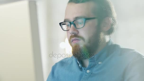 Porträt eines bärtigen jungen Mannes mit Brille, der in seinem Büro sitzt und an einem Computer arbeitet. Computerbildschirm spiegelt sich in seiner Brille. — Stockvideo