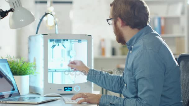 El joven ingeniero barbudo espera un modelo 3D que acaba de crear en su impresora 3D. Se sienta en su escritorio en la oficina moderna . — Vídeos de Stock