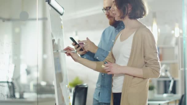 Un jeune homme et une femme discutent du processus de travail sur un tableau blanc. Homme tient Smartphone. Femme dessine sur un tableau blanc . — Video