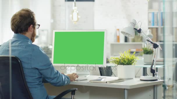 Молода людина з бородою активно працює на своєму комп'ютері. Він сидить у своїй сучасній студії / офісі. Він екран комп'ютера зелений макет . — стокове відео