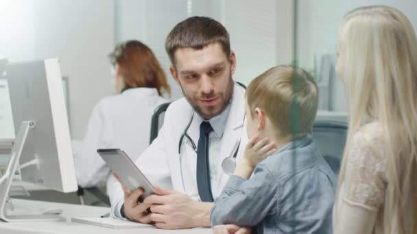 Homme médecin consulte le jeune garçon et sa mère avec l'aide d'une tablette. Ils sourient et plaisantent chaleureusement . — Video