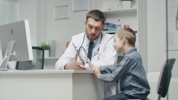 In het ziekenhuis. Knappe glimlachend arts Shows iets op een Tablet-Computer naar een glimlachend kleine jongen en hij beantwoordt met een gebaar. — Stockvideo