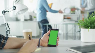 Yakın çekim yeşil ekran Smartphone tutarak ve notlar, yapışkan kağıtlara kalemiyle bir adamın elleri. Arka plan parlak Modern ofiste el sıkışan ve konuşma başlangıç iki erkekle