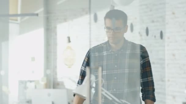 El hombre entra y se sienta en su escritorio, bebe café y comienza a trabajar en una computadora portátil. En segundo plano Hombre trabajando en su computadora de escritorio . — Vídeos de Stock