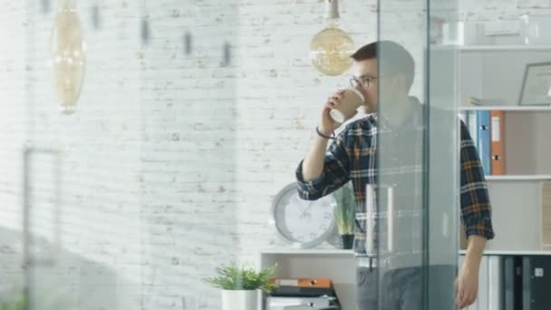 Un homme entre dans un bureau et sirote un café sur son lieu de travail. Commence à travailler sur un ordinateur portable. En arrière-plan Homme travaillant dans sa cabine . — Video