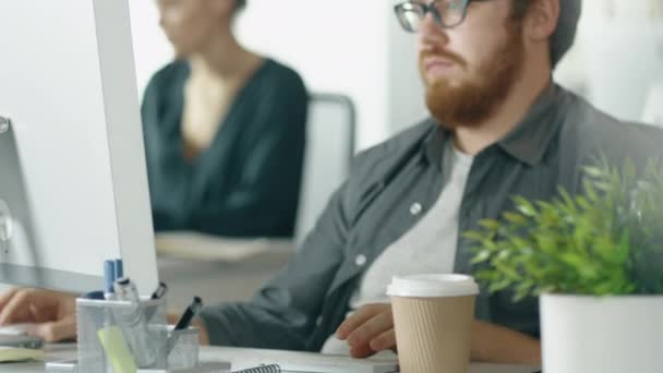 O jovem barbudo trabalha em seu computador desktop, bebe café. No fundo menina trabalha em sua mesa . — Vídeo de Stock