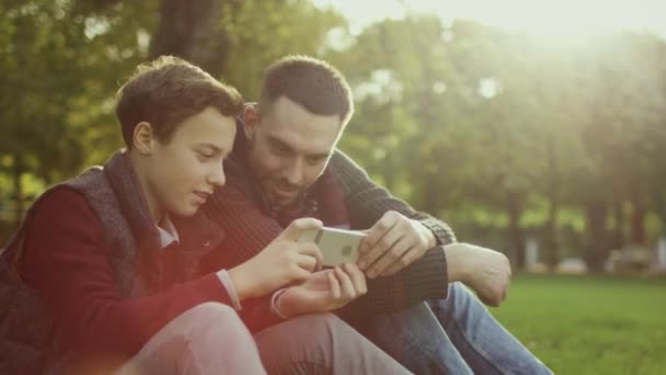 Отец и сын используют смартфон, сидя в парке . — стоковое видео