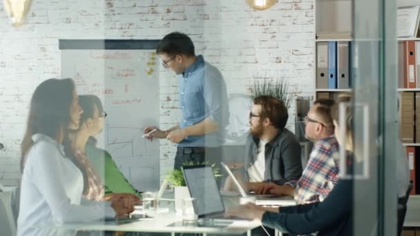 Man rende Whiteboard presentazione al suo staff ufficio creativo. I coworkers si siedono al grande tavolo di vetro con i computer portatili aperti, prendendo appunti. L'ufficio è elegante e luminoso . — Video Stock