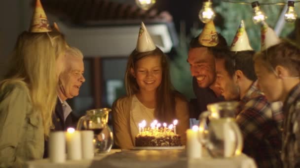 Счастливая улыбающаяся девушка, задувающая свечи в свой праздничный торт. Девушка в окружении семьи и друзей — стоковое видео
