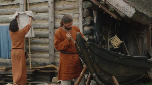 Das Leben der Zivilbevölkerung im Dorf. In mittelalterlicher Kleidung bastelt der Mann ein Boot, während die Frau Kleider aufhängt. Mittelalterliche Reenactment. — Stockvideo