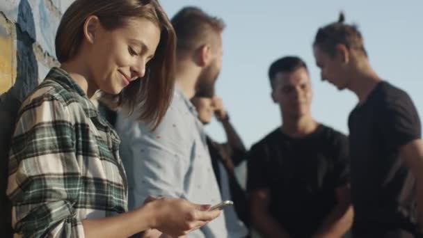 Atractiva adolescente usando el teléfono móvil junto al grupo de sus amigos adolescentes al aire libre en un entorno urbano . — Vídeos de Stock