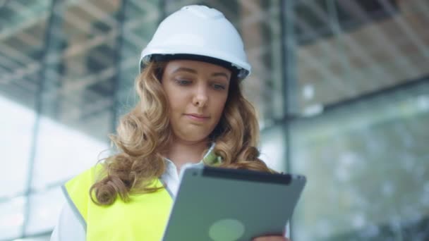 Vrouwelijke ingenieur in Hard Hat en veiligheidsvest met behulp van Tablet PC op de bouwplaats. Glazen gebouw op achtergrond. — Stockvideo